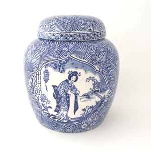 May include: A blue and white ceramic ginger jar with a lid, featuring a detailed illustration of a person in traditional attire holding a fan within a central cartouche, surrounded by a geometric cross-hatch pattern and floral motifs.