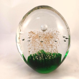 May include: A clear, oval-shaped glass paperweight featuring a solid green base. Suspended within the clear glass are numerous small air bubbles and fine gold-coloured flecks that rise upward from the green section towards the top of the object.