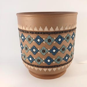 May include: A cylindrical terracotta planter featuring a decorative band of raised blue and light blue diamond-shaped tiles, interspersed with small white and brown floral accents, and bordered by dark brown square tiles and white leaf motifs.