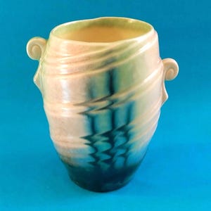May include: A ceramic vase with a distinctive design. The vase has a cream-coloured exterior with swirling patterns of peach, green, and blue. It has two small, curved handles and a dark blue base. The interior is a light yellow colour.