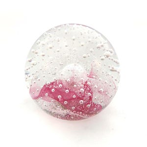 May include: A clear glass paperweight with a pink floral design inside. The round paperweight is filled with small air bubbles, creating a decorative effect. The pink floral element provides a vibrant contrast. A decorative item.