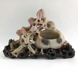 May include: A carved stone sculpture with a central bowl and floral details. The sculpture is light beige with dark brown accents on the base. The floral elements include buds and leaves.