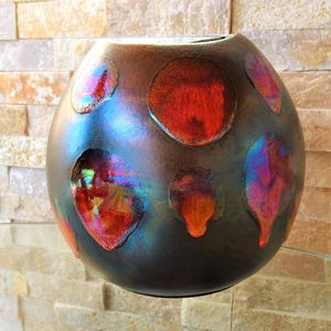 May include: A dark brown, spherical decorative vase with iridescent red, orange, and blue accents. The vase has a textured surface and is displayed against a light-coloured stone wall. The vase is a unique home decor item.