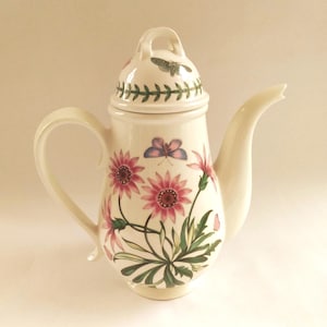 May include: Cream-coloured ceramic coffee pot with a lid, handle, and spout. The pot is decorated with pink and purple flowers, green leaves, and butterflies. The lid has a small handle and a green leaf border.