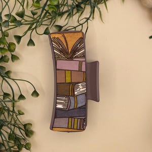 Puede incluir: Una pinza para el pelo de color malva con un diseño de libros apilados en varios colores. La parte superior de la pinza muestra un libro abierto con páginas doradas. Los libros son en tonos de morado, marrón y gris con acentos amarillos.
