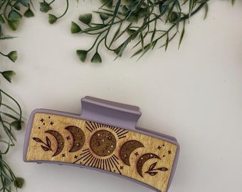 May include: A lavender-colored hair claw clip with a wooden panel featuring a celestial design. The wooden panel displays phases of the moon, a sunburst, and leaf accents. The clip is designed to hold hair in place.