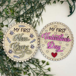 May include: Two wooden, round decorations with scalloped edges. One reads "MY FIRST New Years" in gold and the other reads "MY FIRST Valentines Day" in pink. Both have decorative elements.