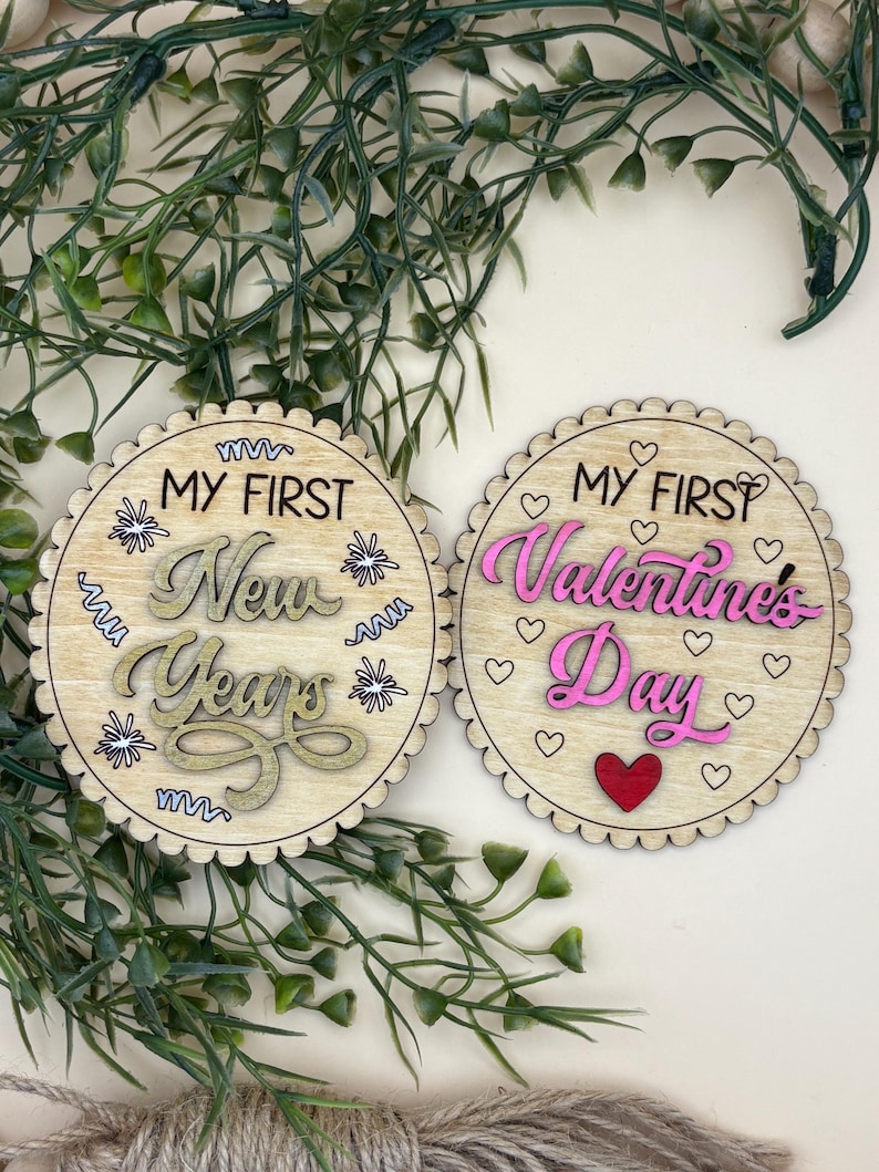 May include: Two wooden, round decorations with scalloped edges. One reads "MY FIRST New Years" in gold and the other reads "MY FIRST Valentines Day" in pink. Both have decorative elements.