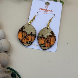 May include: Wooden dangle earrings with a pumpkin design. The earrings feature a light wood grain background with orange pumpkins and green leaves. The earrings are diamond-shaped with gold-colored hooks. The tag reads "PepperJack Designs".