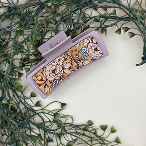 May include: A mauve rectangular hair claw clip with a floral design. The clip features a gold background with beige flowers and blue accents. The clip is surrounded by green foliage and white wooden beads.
