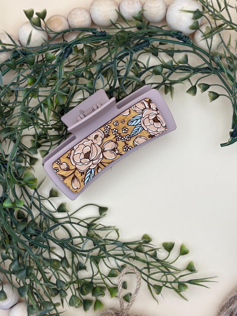 May include: A mauve rectangular hair claw clip with a floral design. The clip features a gold background with beige flowers and blue accents. The clip is surrounded by green foliage and white wooden beads.