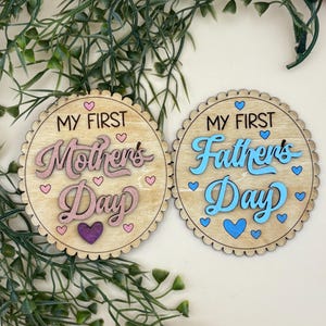May include: Two wooden round decorations with scalloped edges. One reads "My First Mother's Day" in pink script with heart accents. The other says "My First Father's Day" in blue script with heart accents. Greenery and twine are in the background.