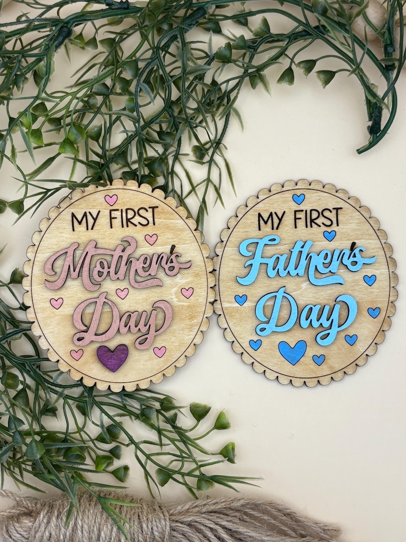 May include: Two wooden round decorations with scalloped edges. One reads "My First Mother's Day" in pink script with heart accents. The other says "My First Father's Day" in blue script with heart accents. Greenery and twine are in the background.