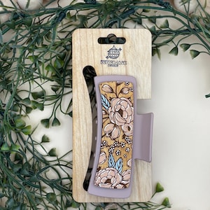 May include: A floral hair claw clip with a light purple base and a wooden accent featuring a beige floral design. The clip is attached to a wooden display card with the text "PepperJack Designs".