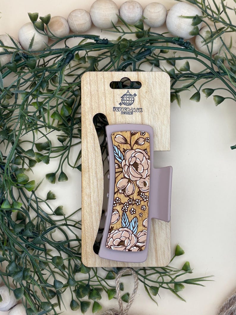 May include: A floral hair claw clip with a light purple base and a wooden accent featuring a beige floral design. The clip is attached to a wooden display card with the text "PepperJack Designs".