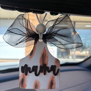 May include: A white car air freshener with a black and brown animal print design. The word "mama" is written in black on the front. The air freshener is hanging from a rearview mirror with a black ribbon.