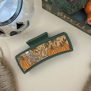 May include: A green rectangular hair claw clip with a wooden design featuring a skeleton, pumpkins, and leaves. The clip has a matte finish and is designed for medium to thick hair. The clip is a Halloween-themed accessory.