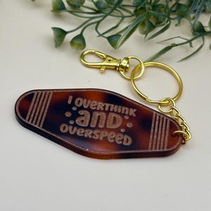 May include: A brown and black keychain with the words "I overthink and overspeed" in a retro motel style. The keychain has a gold-colored clasp and ring.