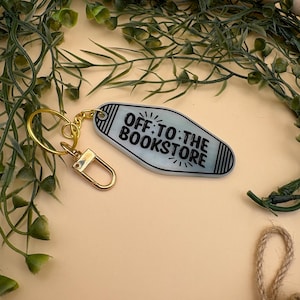 Pode incluir: Um chaveiro azul claro em estilo motel com o texto "OFF TO THE BOOKSTORE" em preto. O chaveiro tem um fecho e um anel dourados. O fundo inclui folhagem verde, contas de madeira e uma superfície bege.