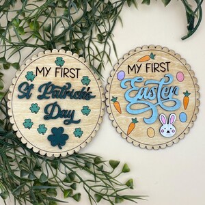 May include: Two wooden round decorations with scalloped edges. One says "My First St. Patrick's Day" with shamrocks. The other says "My First Easter" with carrots, eggs, and a bunny. Both are light brown with green and blue accents.