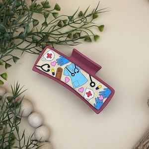 May include: A burgundy hair claw clip with a cream-coloured rectangular design featuring medical-themed illustrations. The design includes a stethoscope, scrubs, a coffee mug, a medical cross, and pink hearts. The clip is surrounded by greenery and wooden beads.