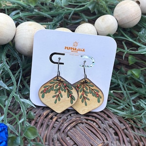 May include: Wooden earrings with a leaf and berry design. The earrings are light brown with a green and red painted design. The earrings are hanging on a white card with the PepperJack Designs logo.