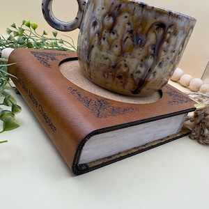 May include: A ceramic mug with a mottled brown, cream, and blue drip glaze sits on a brown book-shaped coaster. The coaster features a circular cutout, decorative black filigree corner patterns, and the text "Just One More Chapter" on the side.