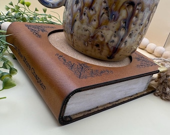 Può includere: Una tazza in ceramica con smalto a goccia marrone, crema e blu posata su un sottobicchiere a forma di libro marrone. Il sottobicchiere presenta un ritaglio circolare, motivi decorativi in filigrana nera agli angoli e il testo "Just One More Chapter".
