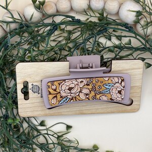 May include: A lavender-colored hair claw clip with a floral design. The clip features a rectangular shape with a wooden base and a clasp. The floral pattern includes beige and pink flowers with blue accents. The clip is displayed on a wooden surface.
