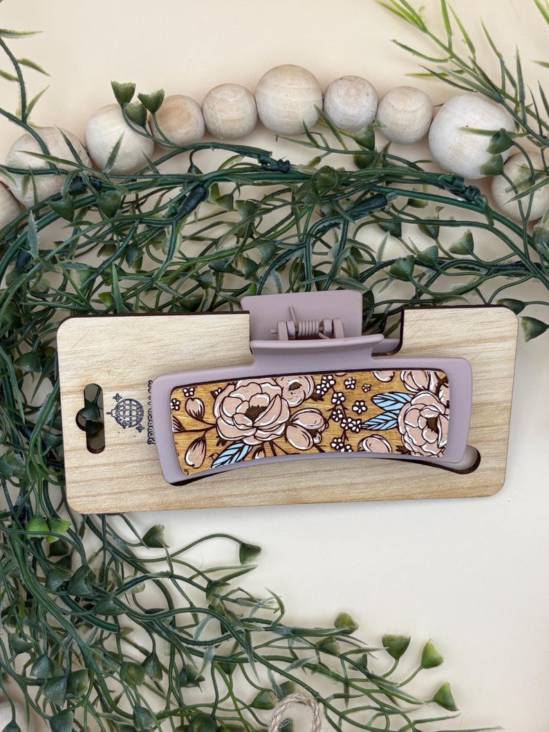 May include: A lavender-colored hair claw clip with a floral design. The clip features a rectangular shape with a wooden base and a clasp. The floral pattern includes beige and pink flowers with blue accents. The clip is displayed on a wooden surface.