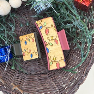 May include: Two wooden hair clips with a Christmas lights design. The clips have a light brown base with colorful light bulb illustrations and a dark pink accent. The clips are set against a woven basket with decorative elements.