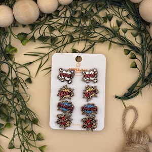 Puede incluir: Un conjunto de seis pendientes de madera estilo cómic en una tarjeta blanca. Los pendientes presentan las palabras "POW", "BANG", "SMASH" y "CRASH" en texto audaz y colorido. La tarjeta está rodeada de vegetación y cuentas de madera.