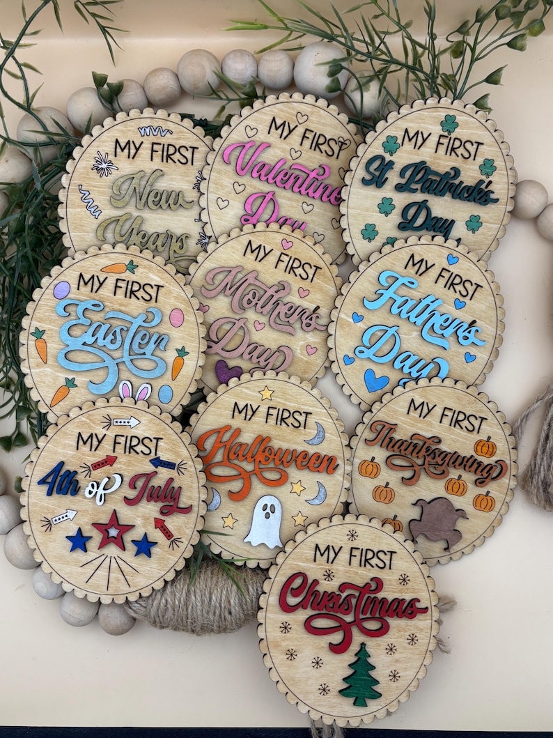 May include: A collection of wooden, circular ornaments with scalloped edges. Each ornament features the phrase "My First" above a holiday name, including New Year's, Valentine's Day, Easter, Mother's Day, Father's Day, 4th of July, Halloween, Thanksgiving, and Christmas.