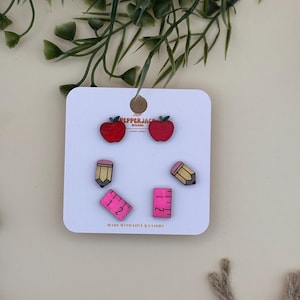 May include: A set of wooden earrings in the shape of apples, pencils, and rulers. The apples are red with green leaves, the pencils are yellow with pink erasers, and the rulers are pink. The earrings are displayed on a white card with the text "MADE WITH LOVE & LASERS".