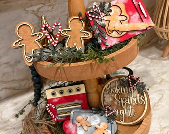 May include: A two-tiered wooden tray decorated with Christmas-themed baking ornaments. The ornaments include gingerbread men, candy canes, a red oven, and a sign that says "Baking Spirits Bright". The tray is filled with greenery.