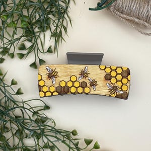 May include: A wooden hair claw clip featuring a bee and honeycomb design. The clip has a gray base and a rectangular wooden top with a yellow honeycomb pattern and several detailed bee illustrations. The clip is a hair accessory.