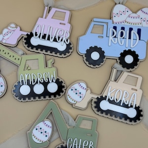 Könnte beinhalten: Oster-Ornamente aus Holz in Form von Baumaschinen, jeweils mit einem Namen. Die Fahrzeuge sind in Pastellfarben wie Rosa, Blau und Grün lackiert, mit schwarz-weißen Rädern. Jedes Ornament hat ein Band zum Aufhängen.