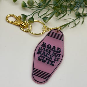 Puede incluir: Un llavero rosa estilo motel con el texto "ROAD RAGE BUT MAKE IT CUTE" en negro. El llavero tiene un cierre y un anillo de color dorado.