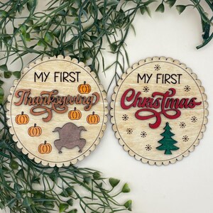 May include: Two wooden round ornaments with scalloped edges. One says "MY FIRST Thanksgiving" with pumpkins and a turkey. The other says "MY FIRST Christmas" with snowflakes and a Christmas tree.