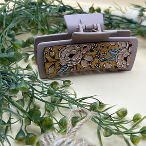 May include: A light purple hair claw clip with a floral design. The clip features a rectangular wooden panel with a pattern of tan and white flowers with blue accents. The clip is made of plastic and has a spring mechanism.