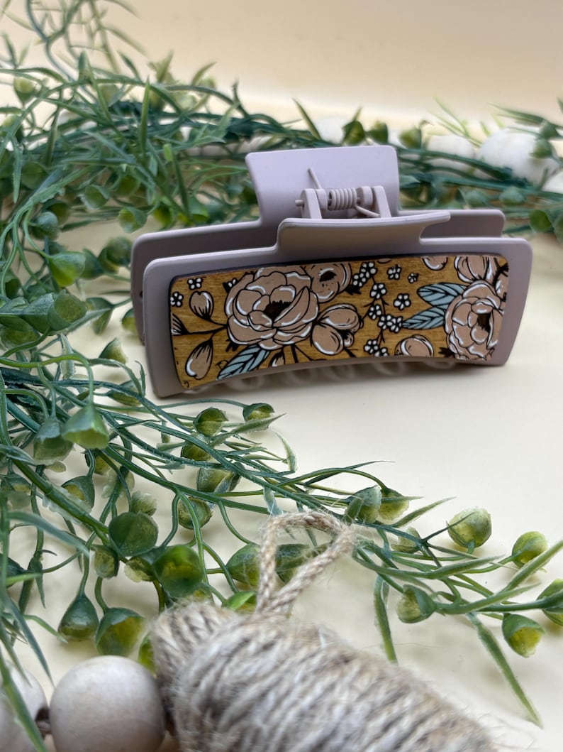 May include: A light purple hair claw clip with a floral design. The clip features a rectangular wooden panel with a pattern of tan and white flowers with blue accents. The clip is made of plastic and has a spring mechanism.