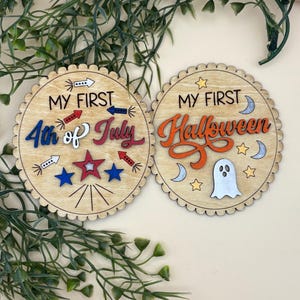 May include: Two wooden, scalloped-edge coasters. One says "MY FIRST 4th of July" with red, white, and blue accents. The other says "MY FIRST Halloween" with orange, white, and silver accents. Decorative items.