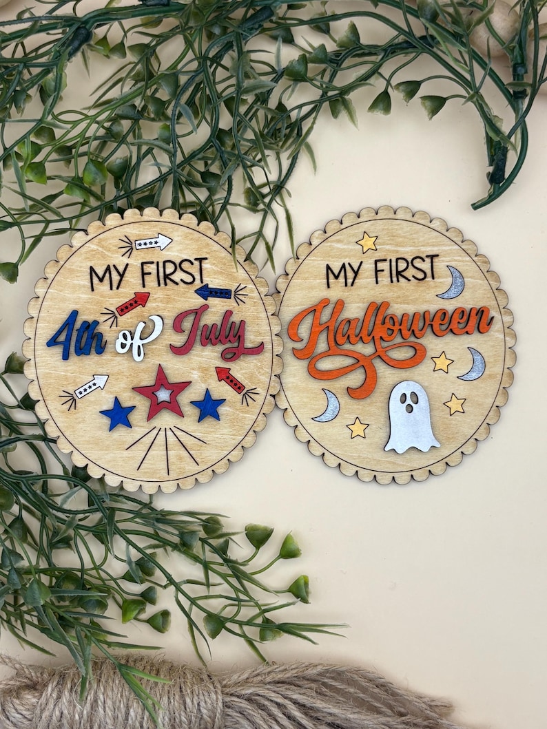 May include: Two wooden, scalloped-edge coasters. One says "MY FIRST 4th of July" with red, white, and blue accents. The other says "MY FIRST Halloween" with orange, white, and silver accents. Decorative items.
