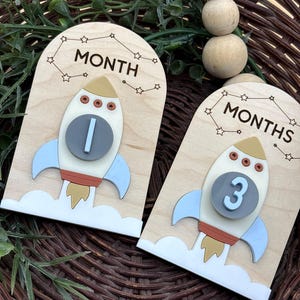 May include: Two wooden milestone plaques with rocket ship designs. One plaque reads "MONTH" with a rocket ship displaying the number "1". The other reads "MONTHS" with a rocket ship displaying the number "3". The rockets are white, blue, and tan.