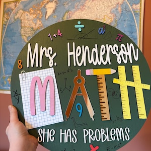 May include: A circular, green wooden sign with the text "Mrs. Henderson MATH SHE HAS PROBLEMS." The sign features math symbols, a pencil, a protractor, and the number 9. The letters "M" and "A" are pink and copper, respectively.