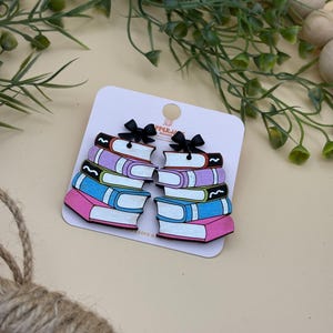 May include: Wooden book-shaped earrings with pink, blue, purple, and white pages. Each earring has a stack of books with a black bow at the top. The earrings are on a white card with the text "PPER.J" and "WITH LOVE &" printed on it.