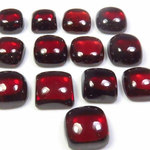 Könnte beinhalten: Eine Ansammlung von dreizehn tiefroten, quadratischen Cabochon-Edelsteinen mit abgerundeten Ecken und glatten, polierten Oberflächen. Jeder Stein hat ein hochglänzendes Finish, das Licht reflektiert und helle weiße Glanzlichter auf der dunkelroten Oberfläche erzeugt.
