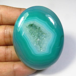 May include: Oval-shaped polished agate stone in shades of teal and white. The stone features a central geode cavity filled with small, sparkling crystals. The agate has a smooth, glossy surface and is held by a hand.