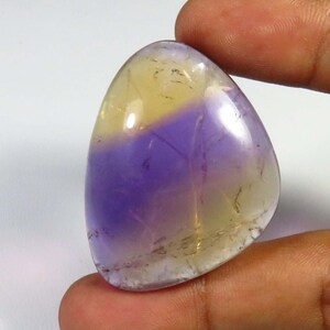 May include: A polished gemstone with a smooth, rounded shape. The stone features a gradient of colors, with a yellow-gold upper half and a deep purple lower half. The gemstone is held between fingers, showcasing its size and luster.