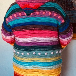 Puede incluir: Un c&aacute;rdigan de crochet a rayas de colores con capucha. El c&aacute;rdigan presenta un esquema de color arco&iacute;ris con rayas rosas, moradas, azules, verdes, amarillas, naranjas y rojas. La capucha est&aacute; forrada de rosa y tiene un ribete de crochet blanco.